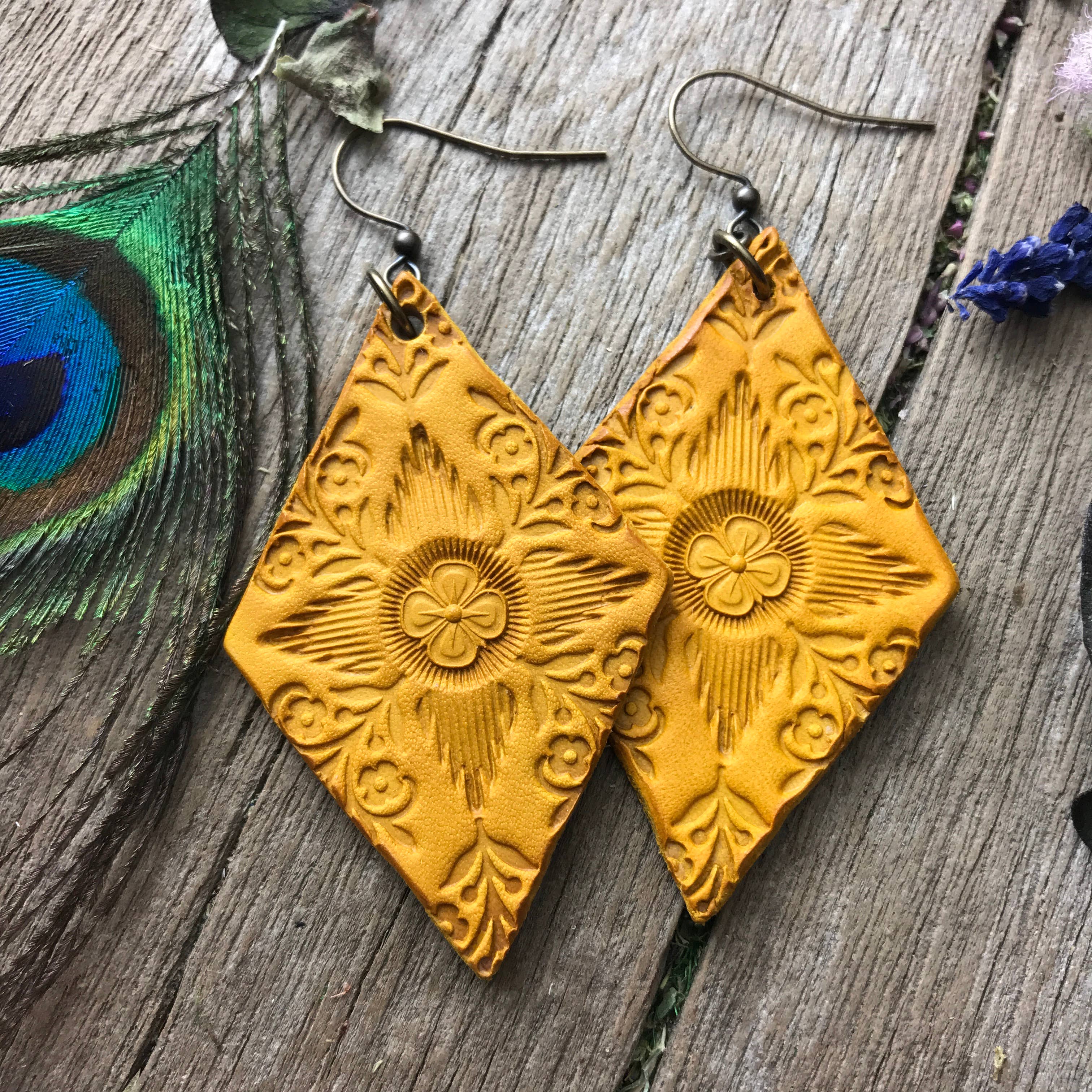 Pair of yellow hand tooled leather earrings on a wooden surface with a peacock feather and lavender flowers.