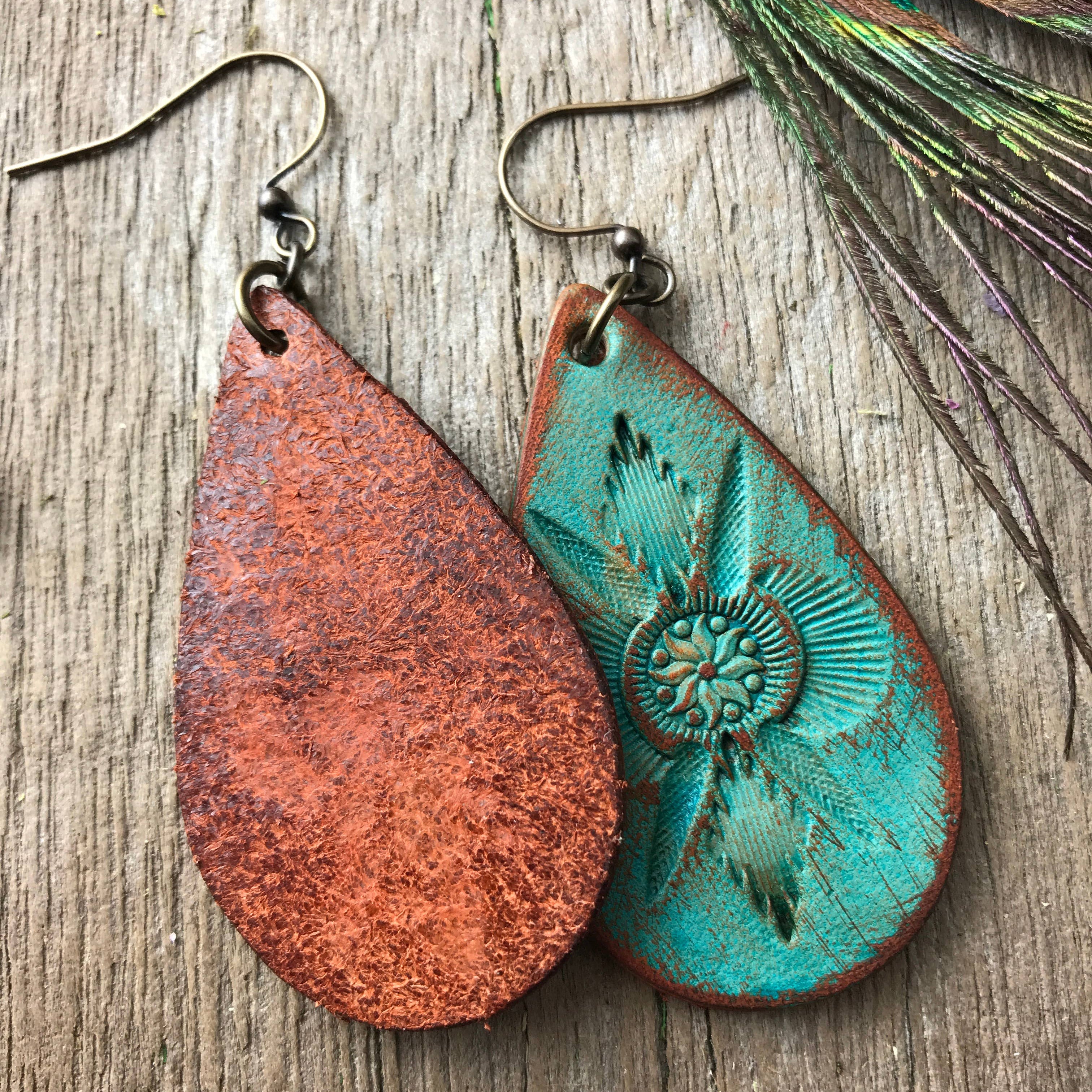 Teardrop-shaped leather earrings with one flipped to the back side and the other front side up on a wooden surface with peacock feathers.