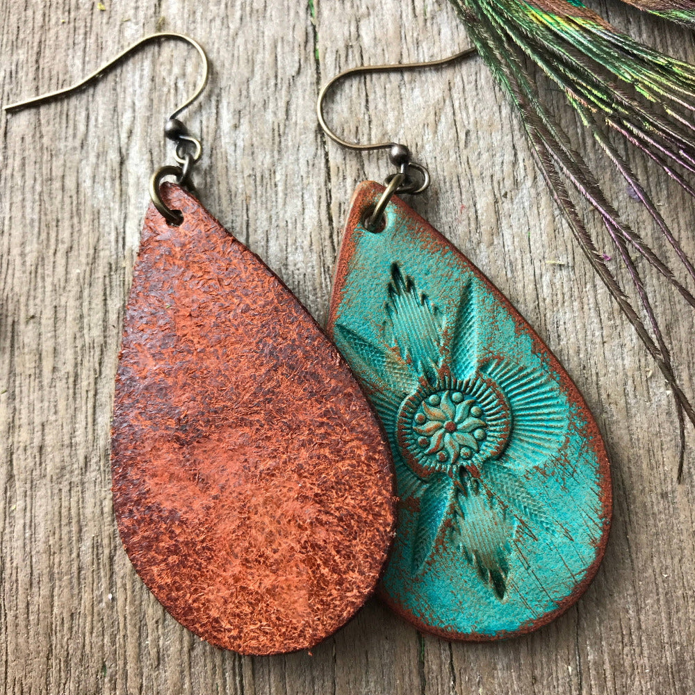 Teardrop-shaped leather earrings with one flipped to the back side and the other front side up on a wooden surface with peacock feathers. Handmade in USA
