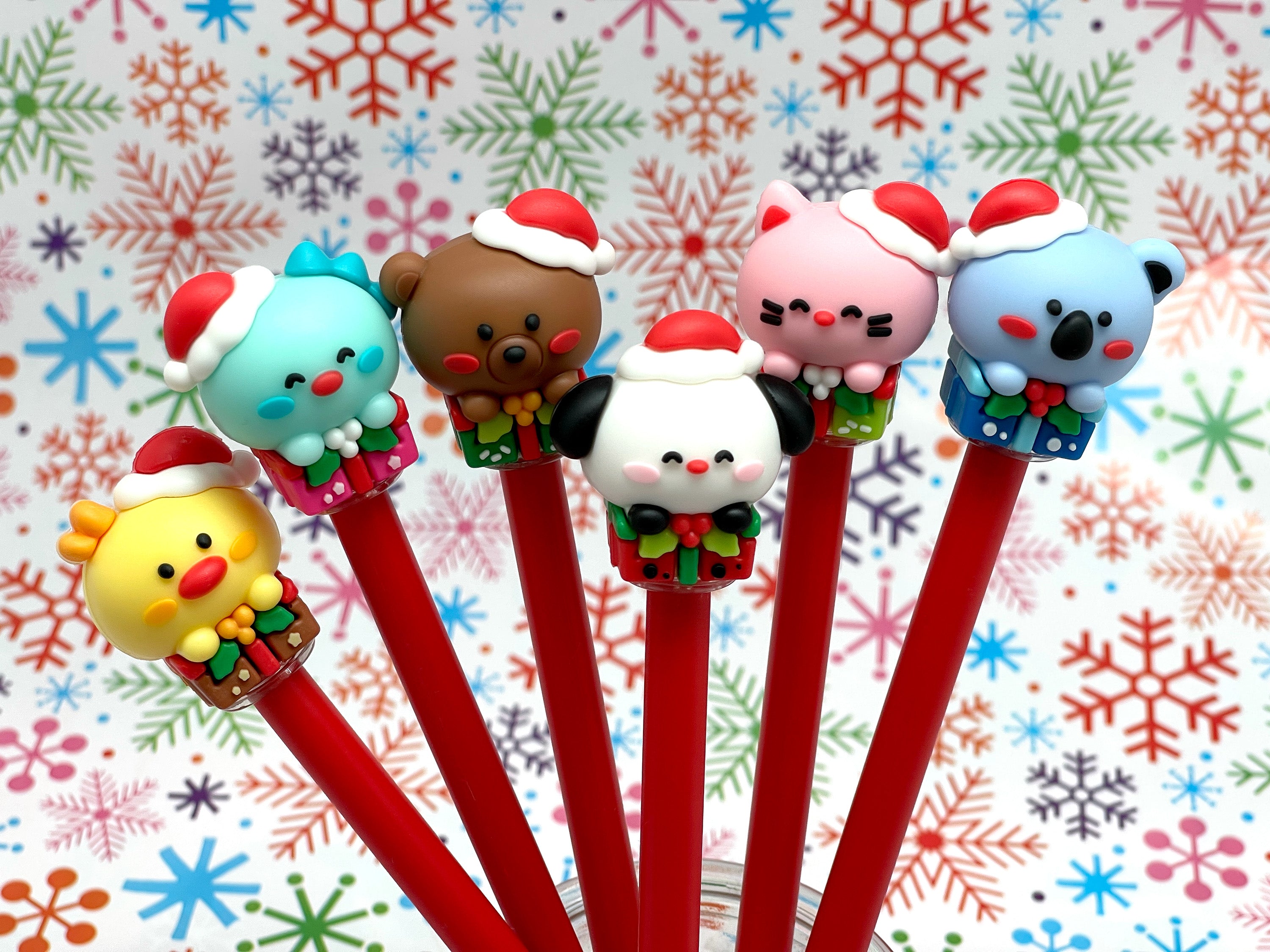 Set of six gel pens with Kawaii style animals wearing Christmas hats and sitting on gift boxes on the pen lid with a colorful snowflake background.