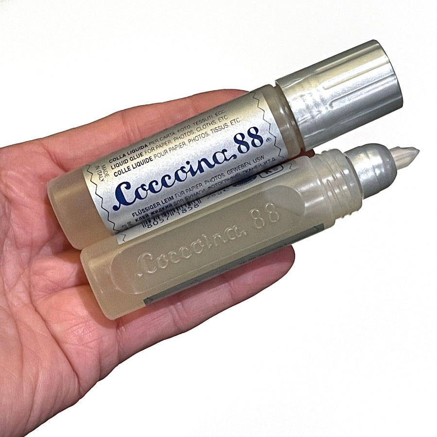 Adult hand holding 2 tubes of Coccoina liquid glue against a white background