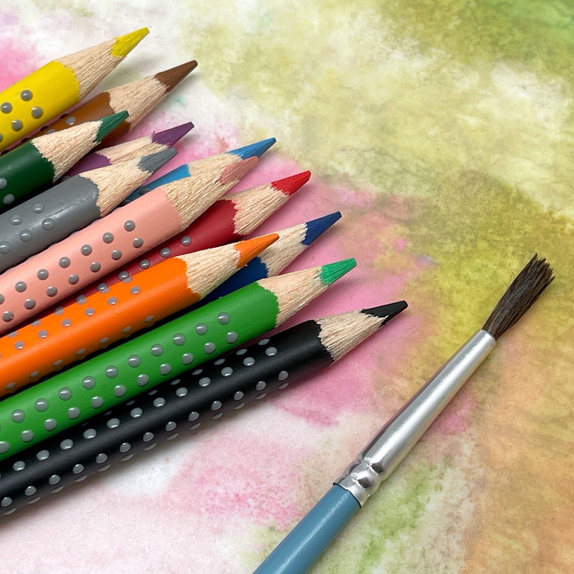 A set of grip watercolor pencils in various colors, each featuring a unique ergonomic triangular design with soft dots for grip and a paint brush on a colorful background.