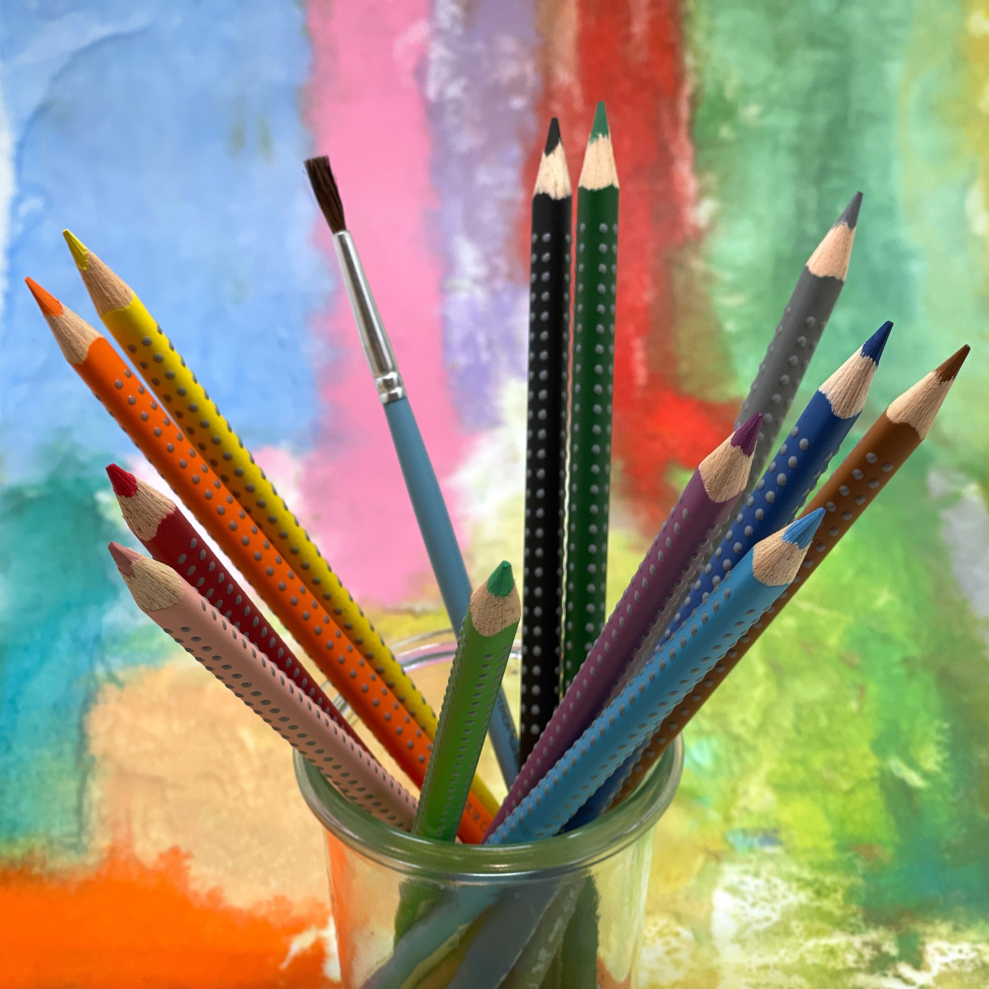 Colorful watercolor pencils and a paintbrush in a glass jar against a colorful abstract background