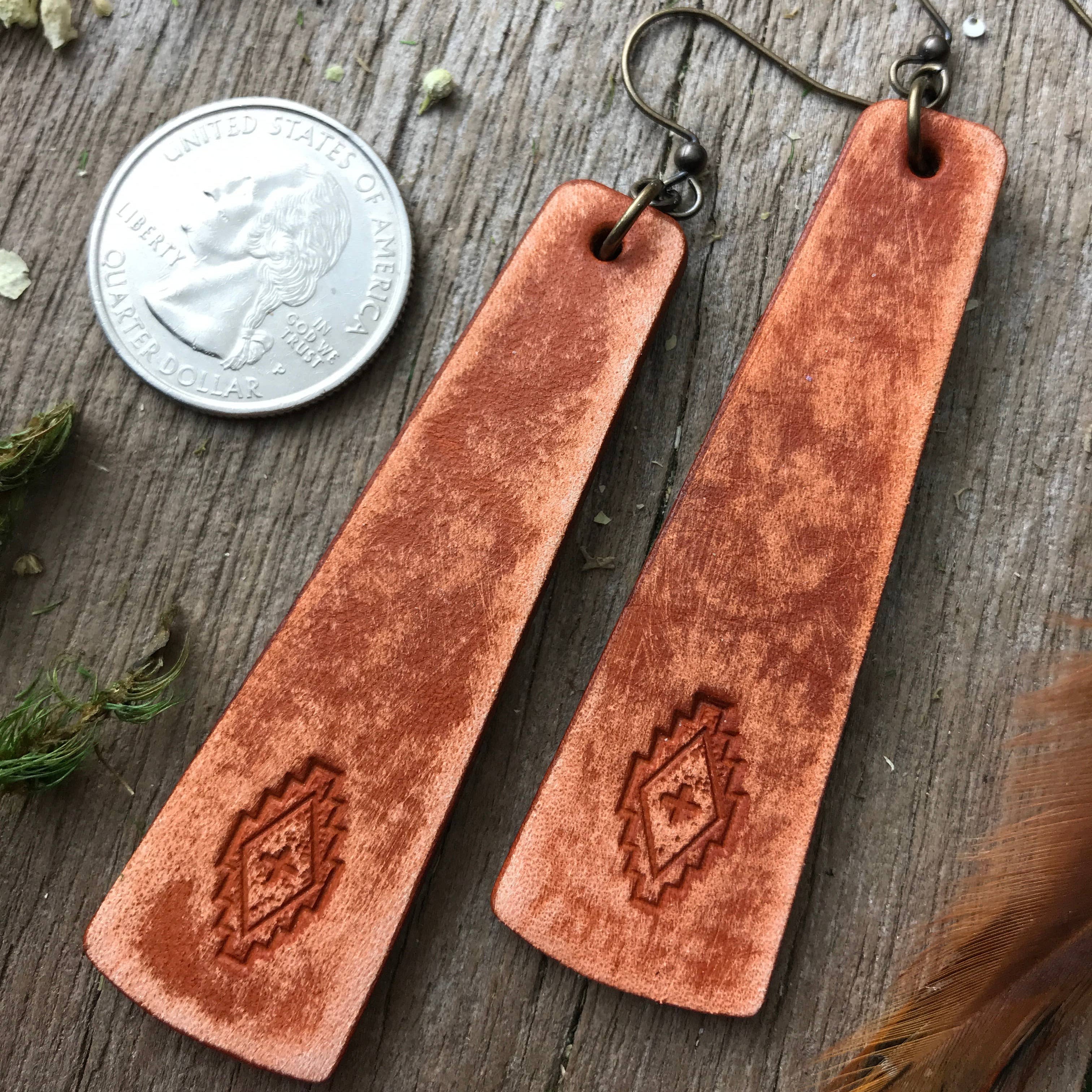Brown leather earrings on a wooden surface with a coin for scale.