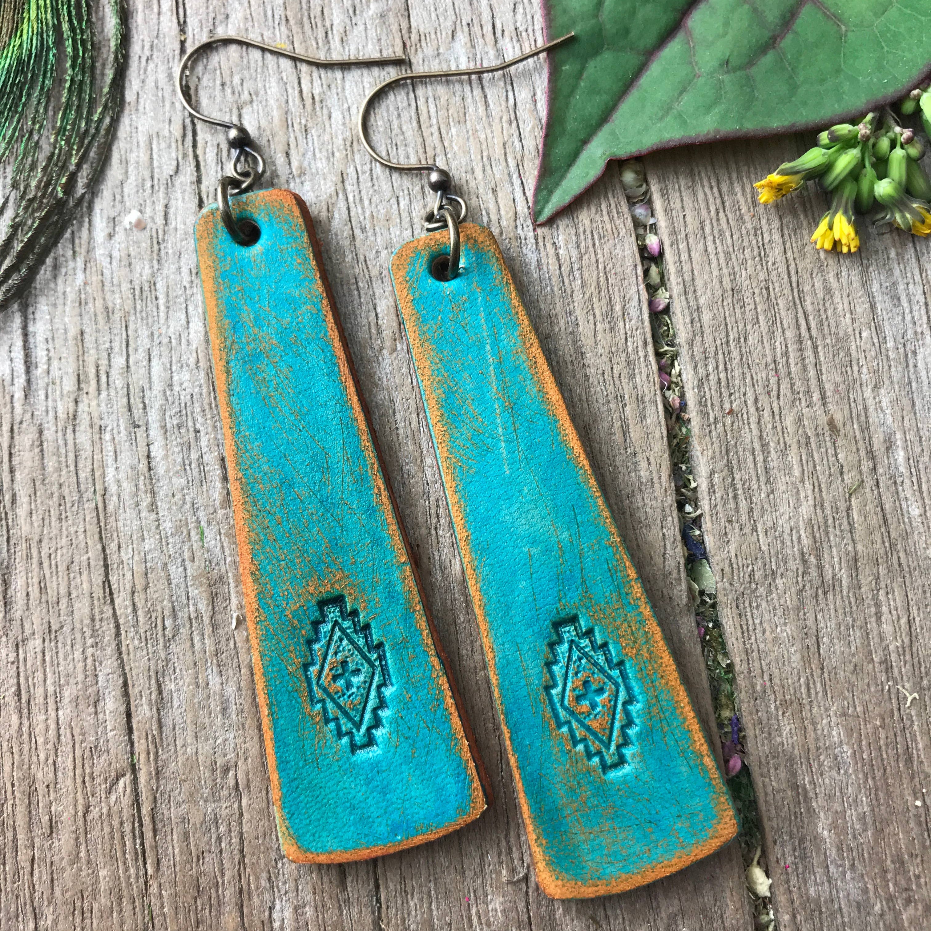 Turquoise leather earrings with brown accents on a wooden surface with green leaves.
