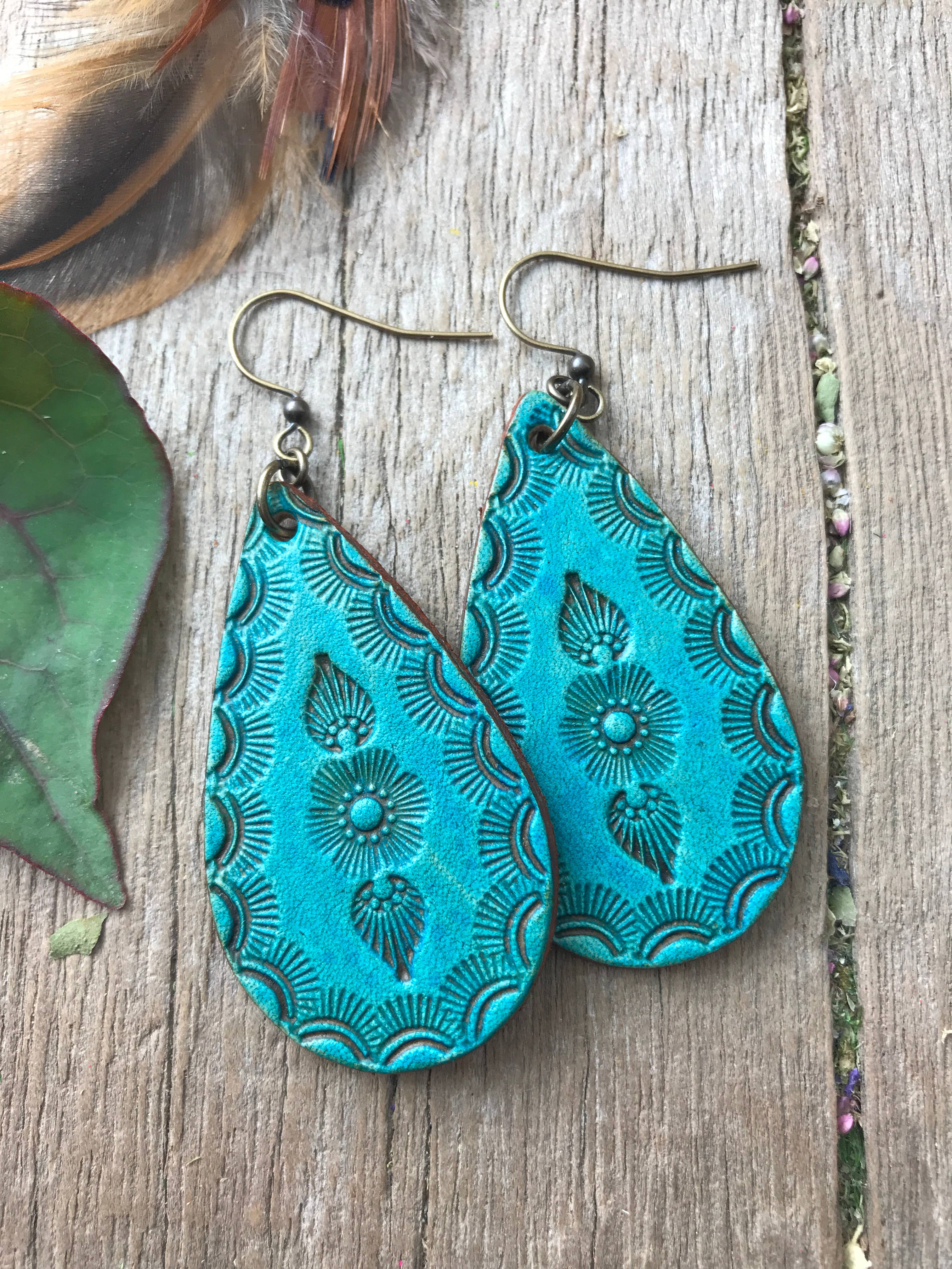 Turquoise embossed leather earrings on a wooden surface with leaves and feathers.
