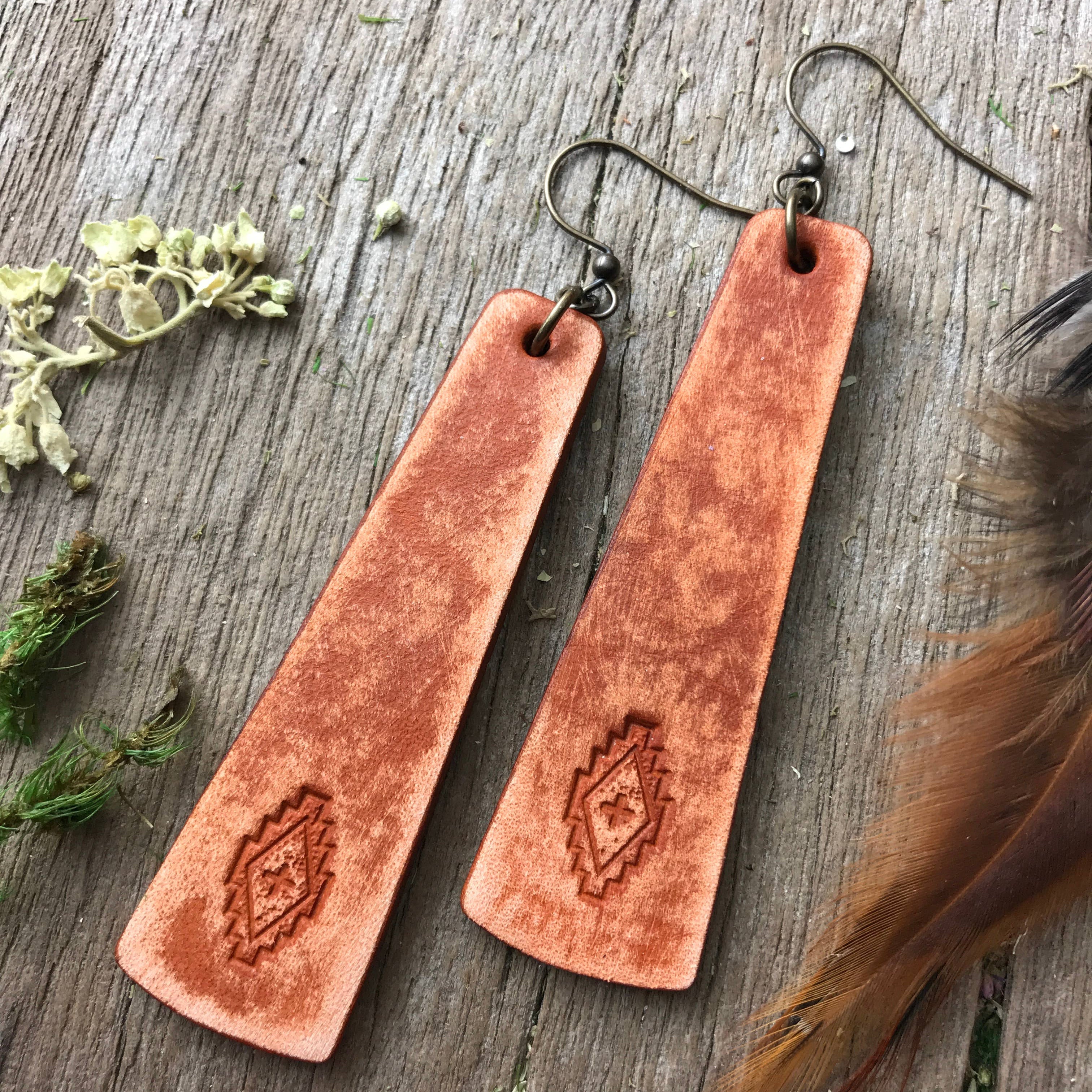 Pair of brown leather earrings with embossed design on a wooden surface with feathers and greenery.