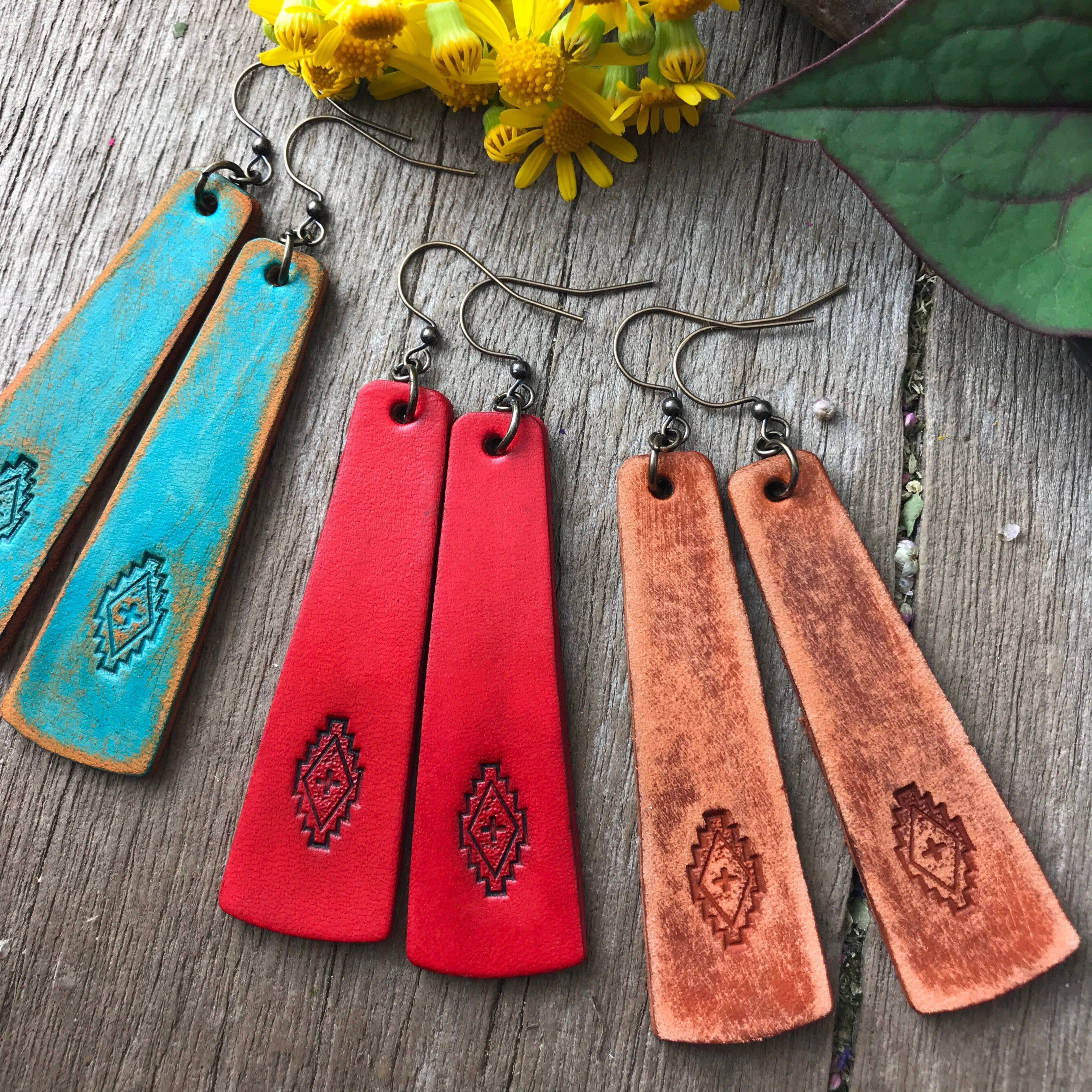 Three pairs of leather earrings in blue, red, and brown on a wooden surface with flowers and leaves.