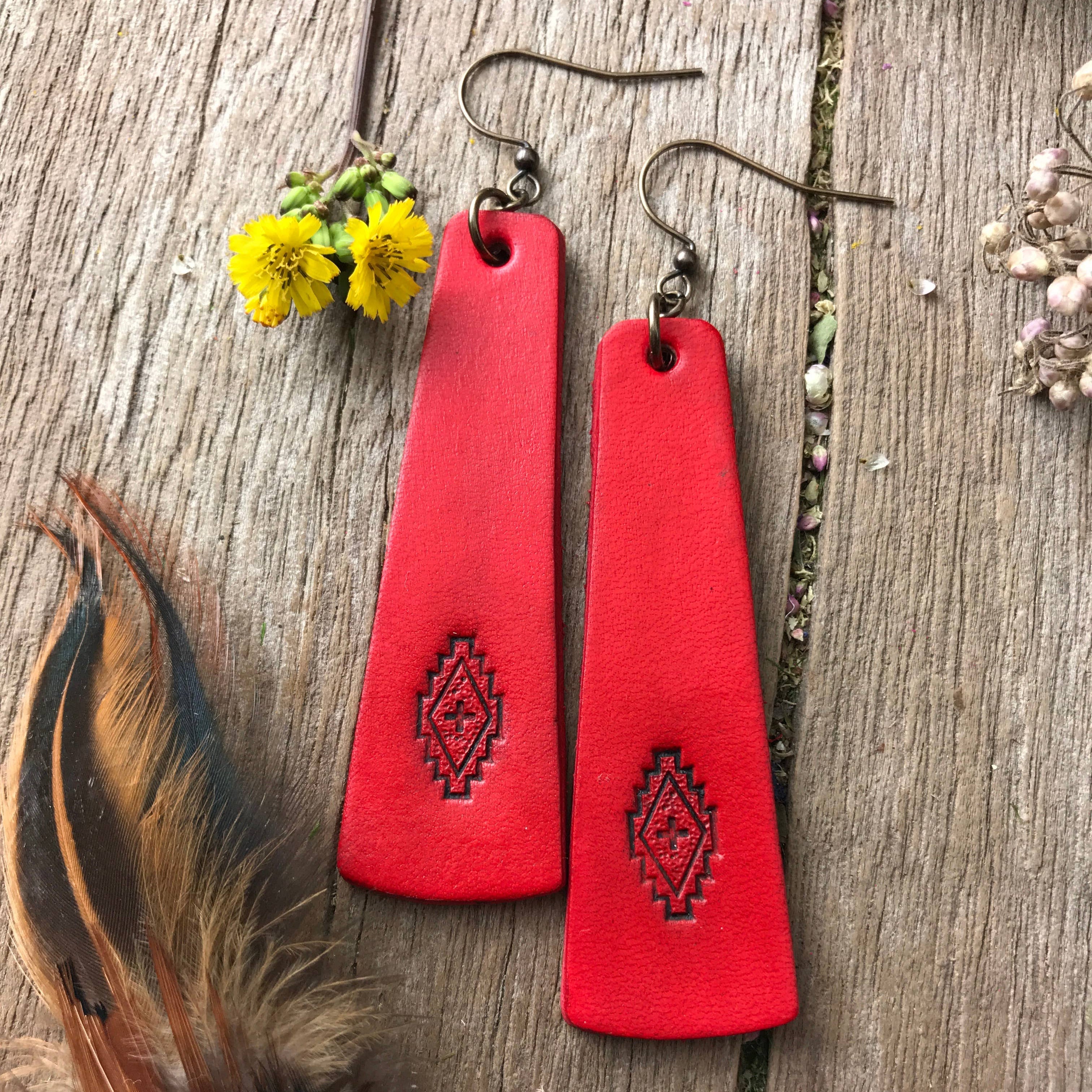 Red leather earrings with tribal design on a wooden surface with feathers and flowers.