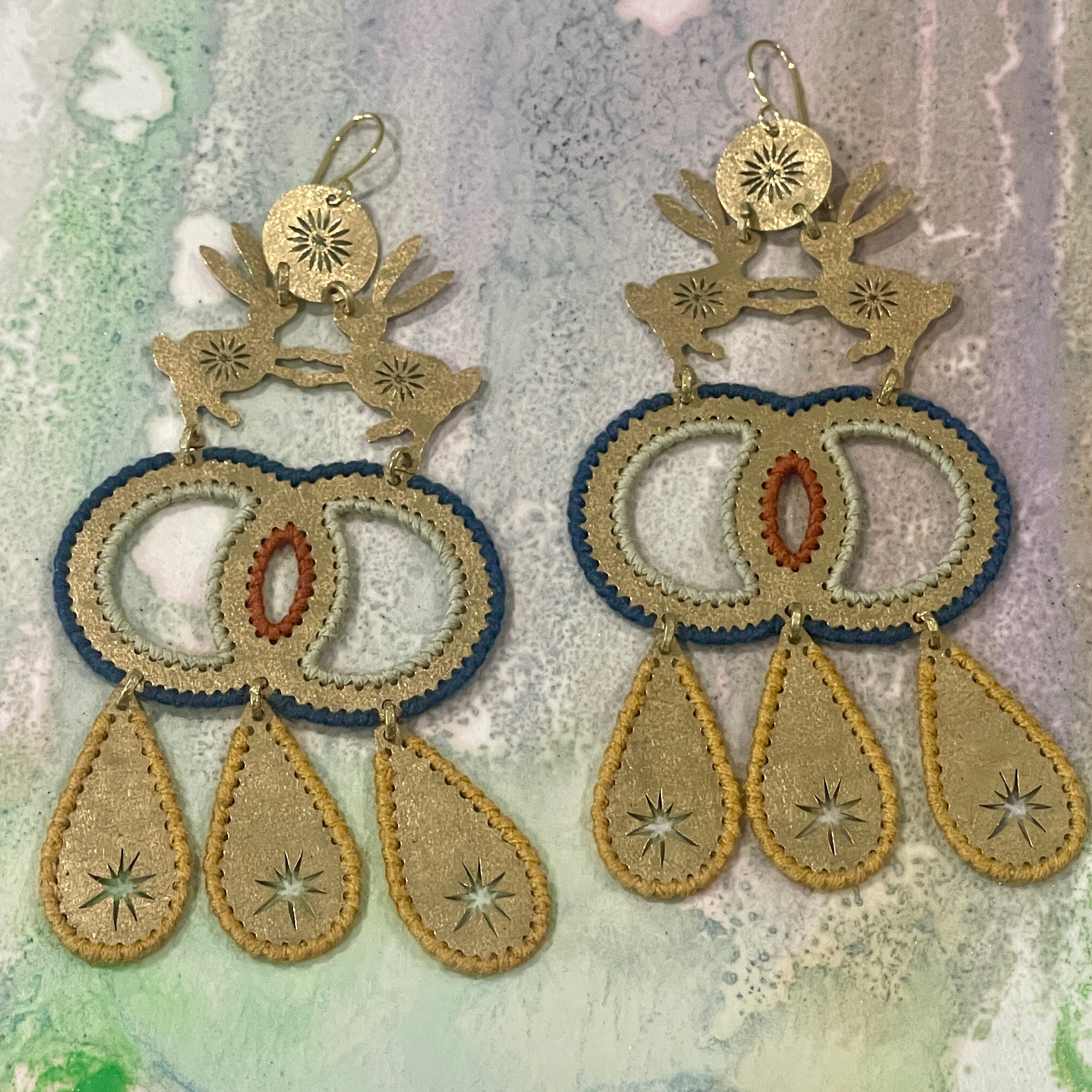 Pair of dangle earrings made from textured brass with two hares dancing together and color thread edges on a muted pastel color background