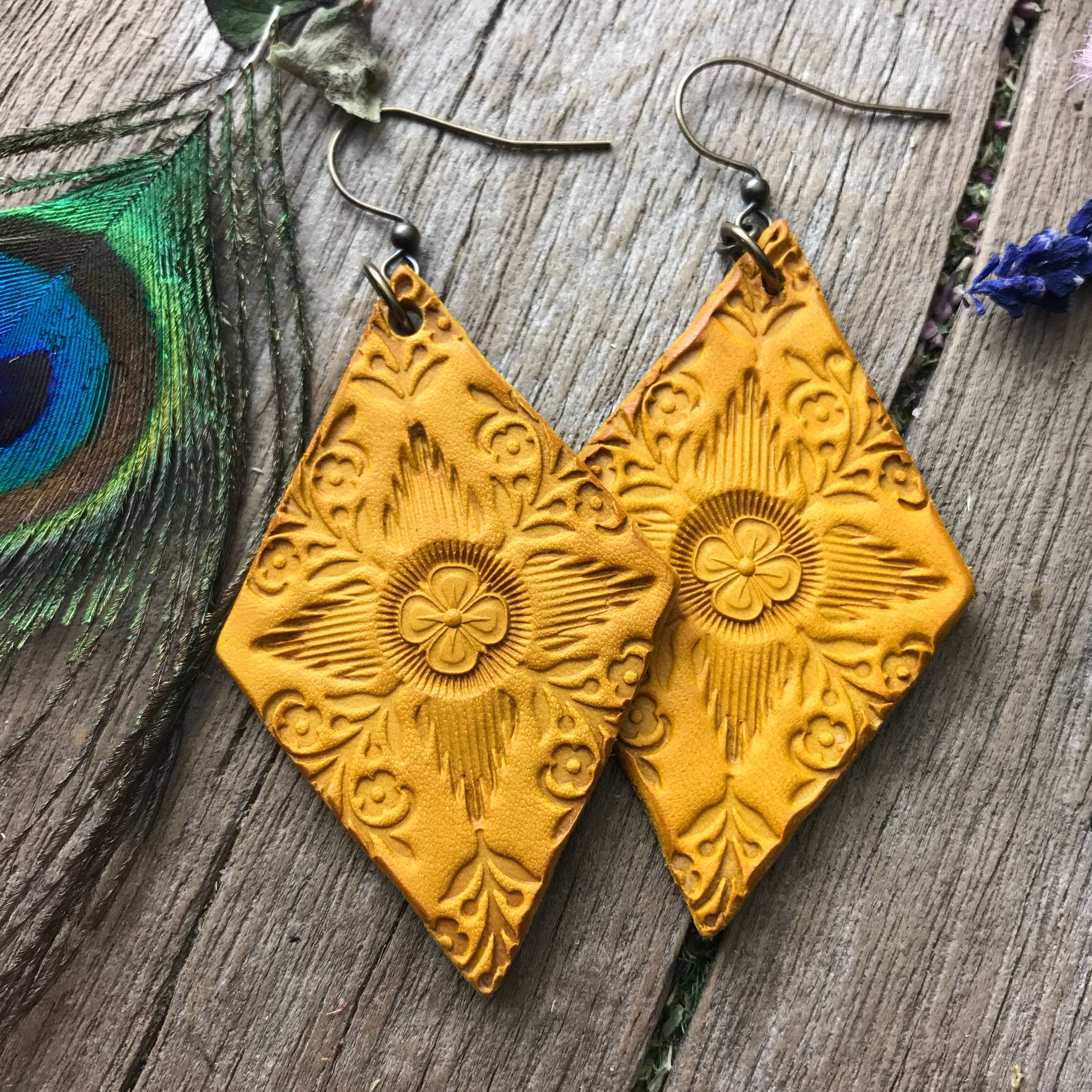 Pair of yellow hand tooled leather dangle boho earrings on a wooden surface with a peacock feather and lavender flowers.