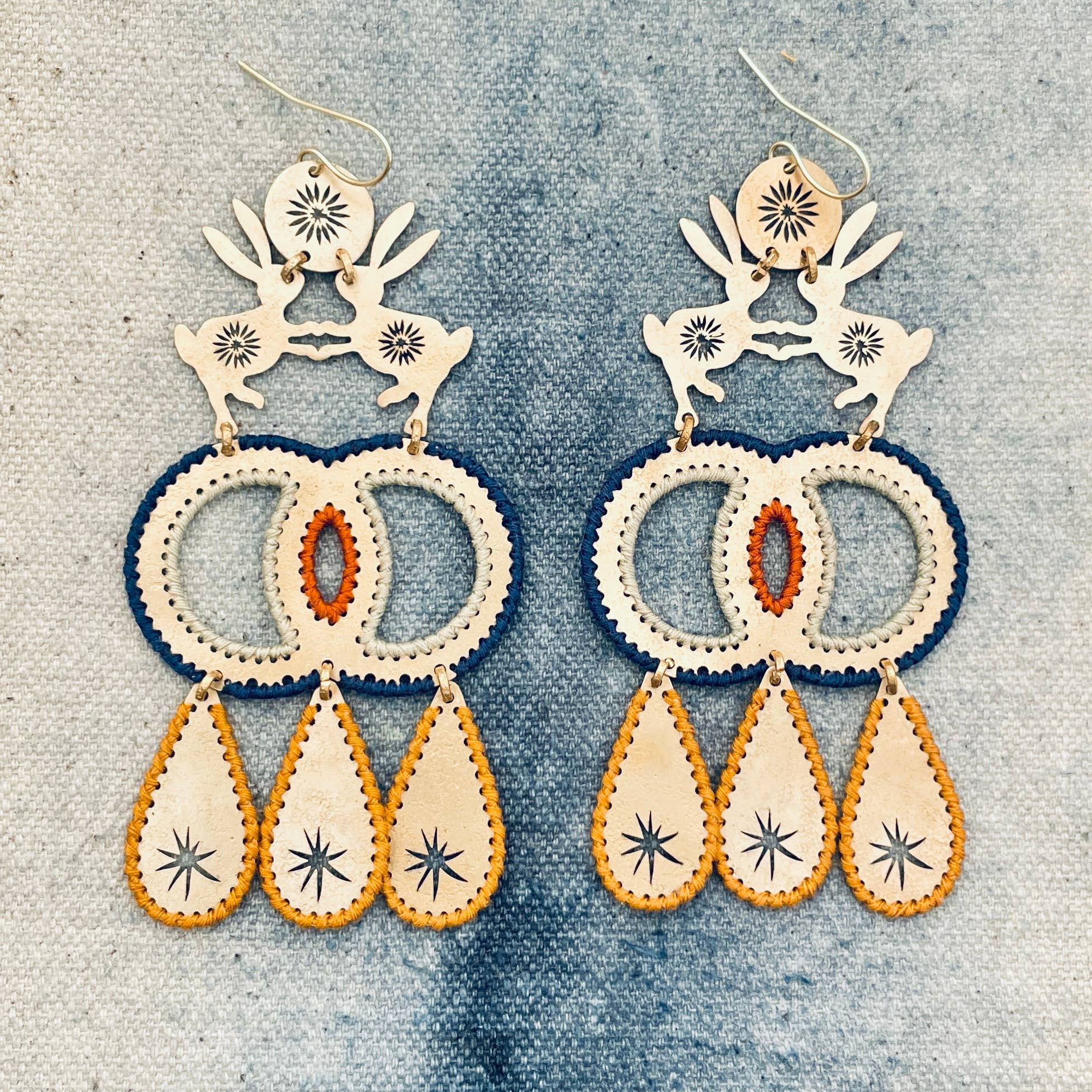 Pair of handmade brass dangle earrings in boho chic style with intricate designs of two hares dancing and hand stitched thread edges on a textured fabric background