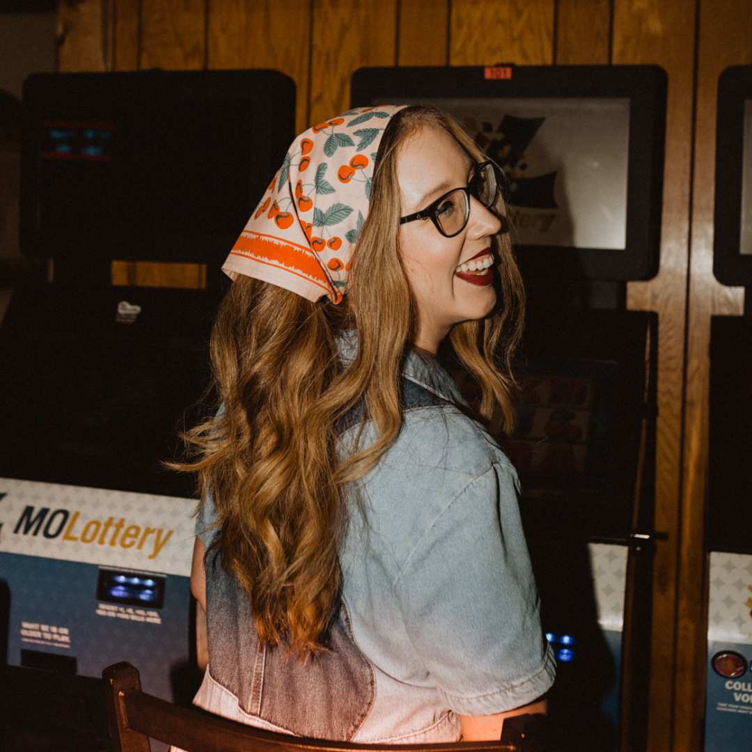 Woman with a cherry pattern bandana and glasses smiling in front of a slot machine.