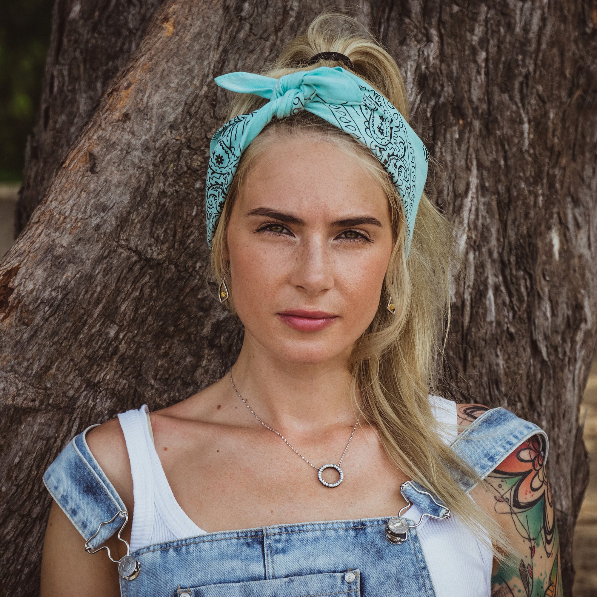 Woman artist wearing cotton bandana headband and denim overalls standing against a tree.