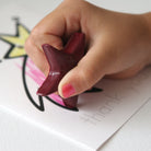 Child's hand holding a red star-shaped crayon over a piece of paper with writing and drawings.