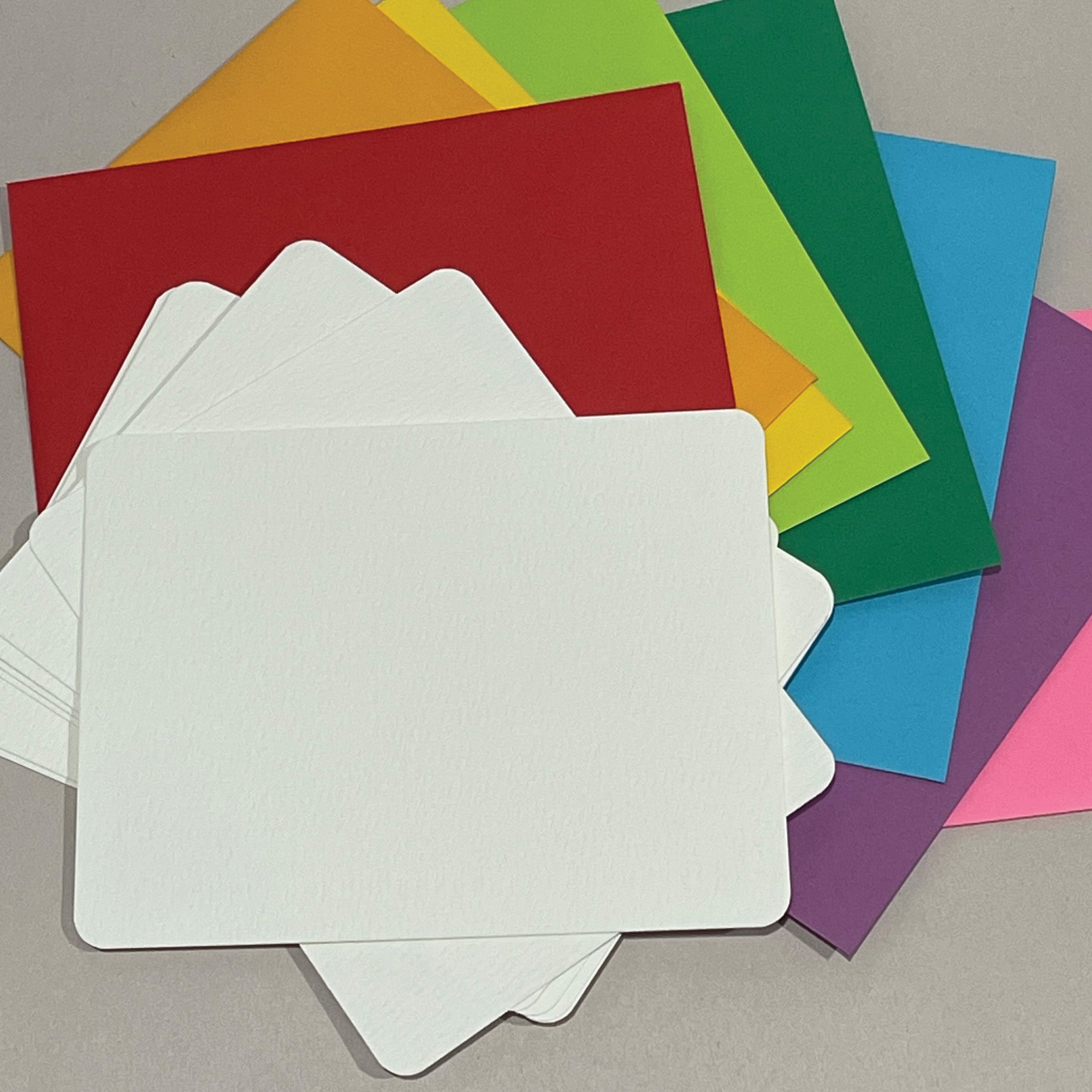A stack of blank watercolor paper flat cards with rounded corners on top of a stack of colorful A2 envelopes