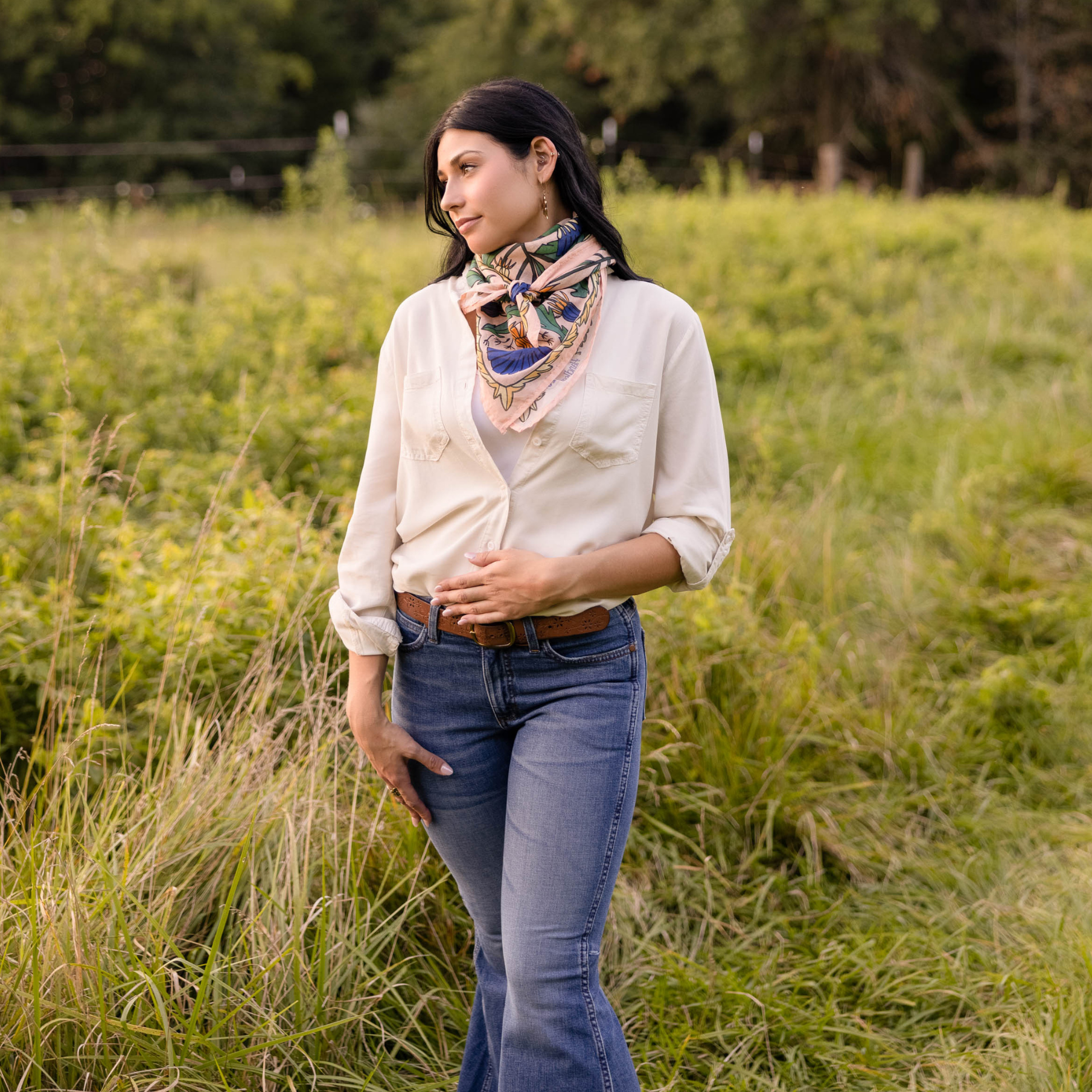 Woman standing in a grassy field wearing a white shirt, blue jeans, and a patterned wild rag scarf.