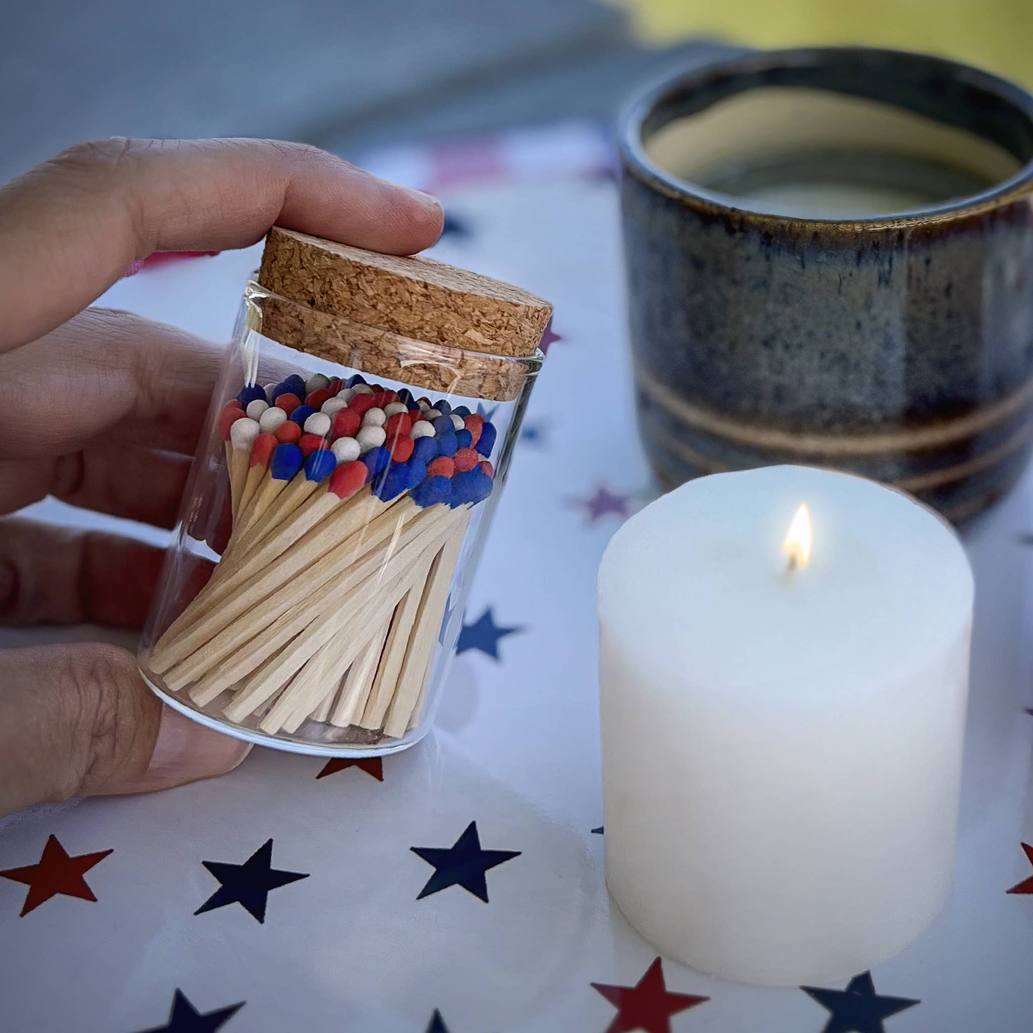 Hand holding a glass container with matches and colorful beads, next to a lit candle on a star-patterned surface.