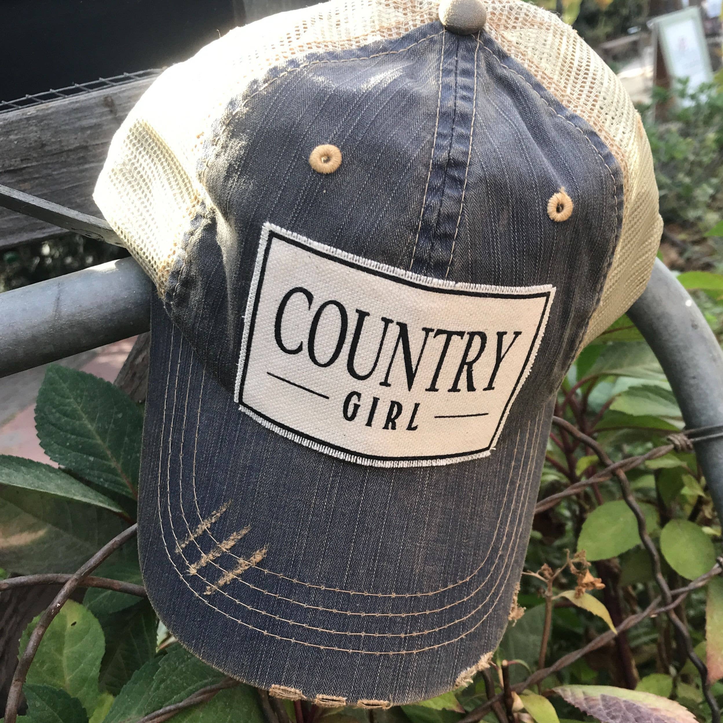 Trucker hat with 'Country Girl' patch on a metal railing with greenery in the background