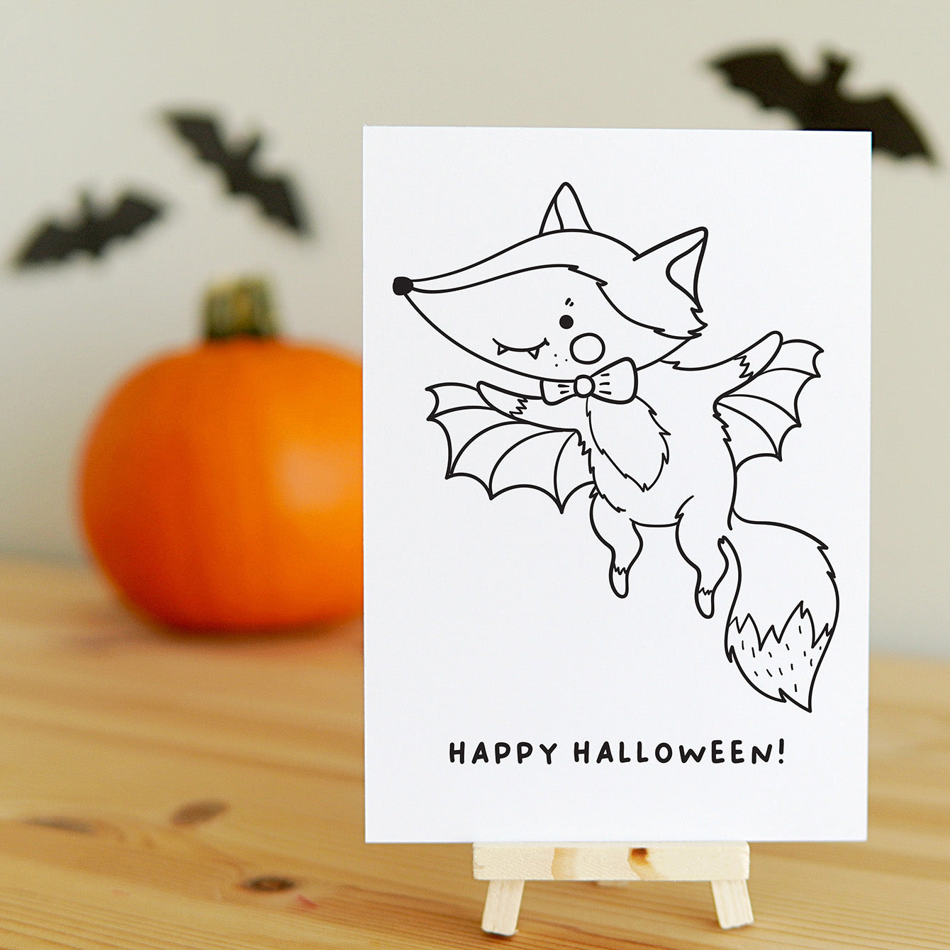 Halloween-themed card with a fox dressed as a vampire on a wooden surface with a pumpkin and bats in the background.