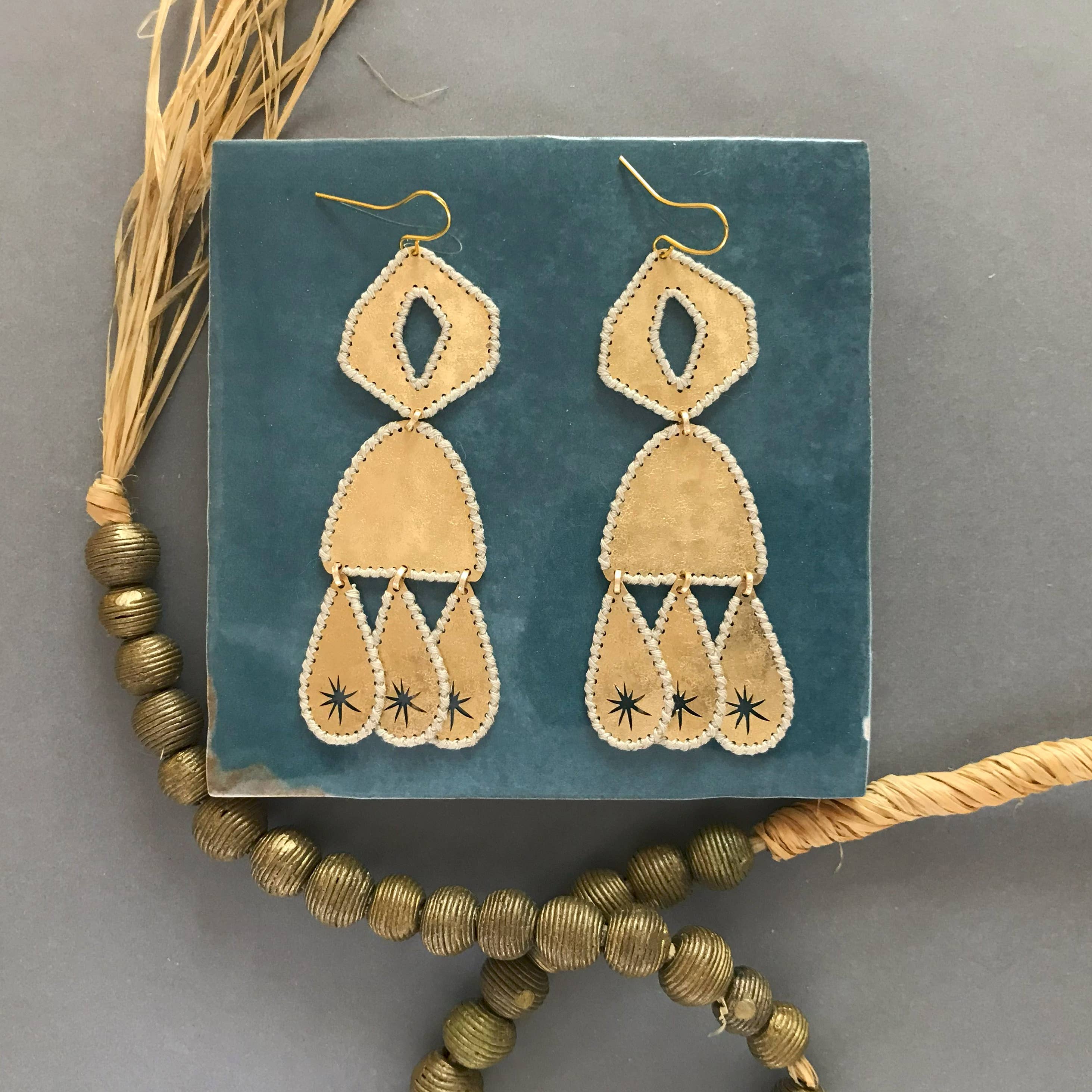 Pair of gold-tone brass handmade earrings with stitched thread edges on a teal card with decorative elements