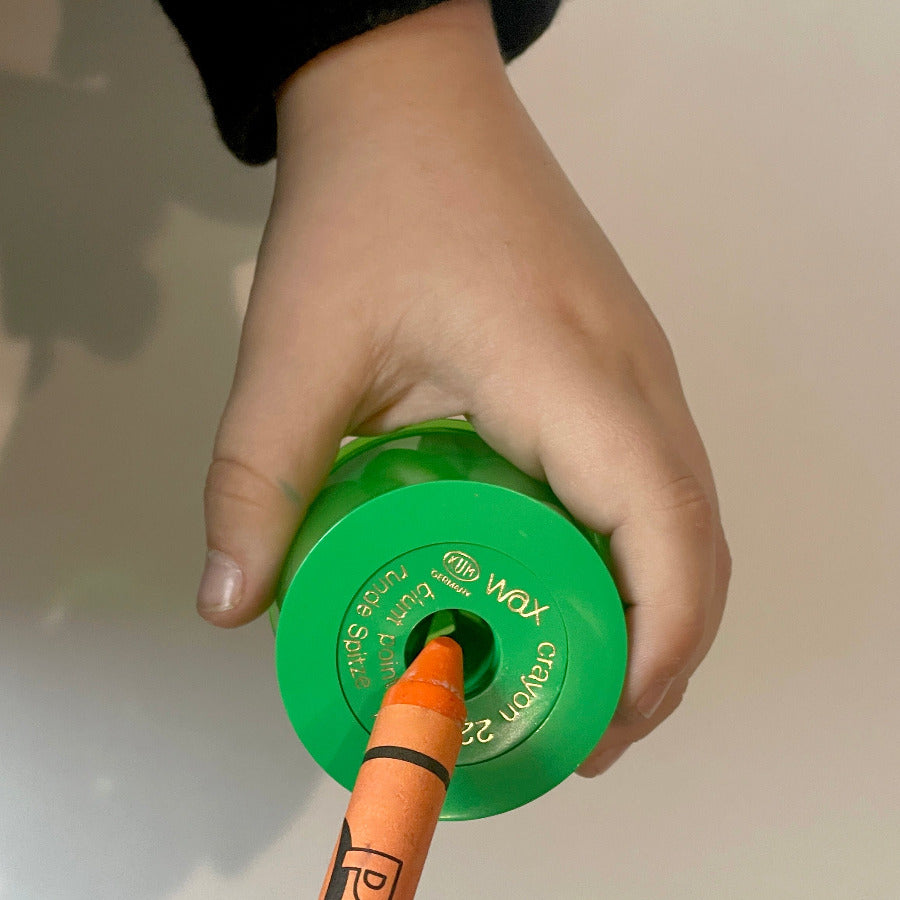 Child hand holding a green wax crayon sharpener and an orange jumbo crayon against a cream background