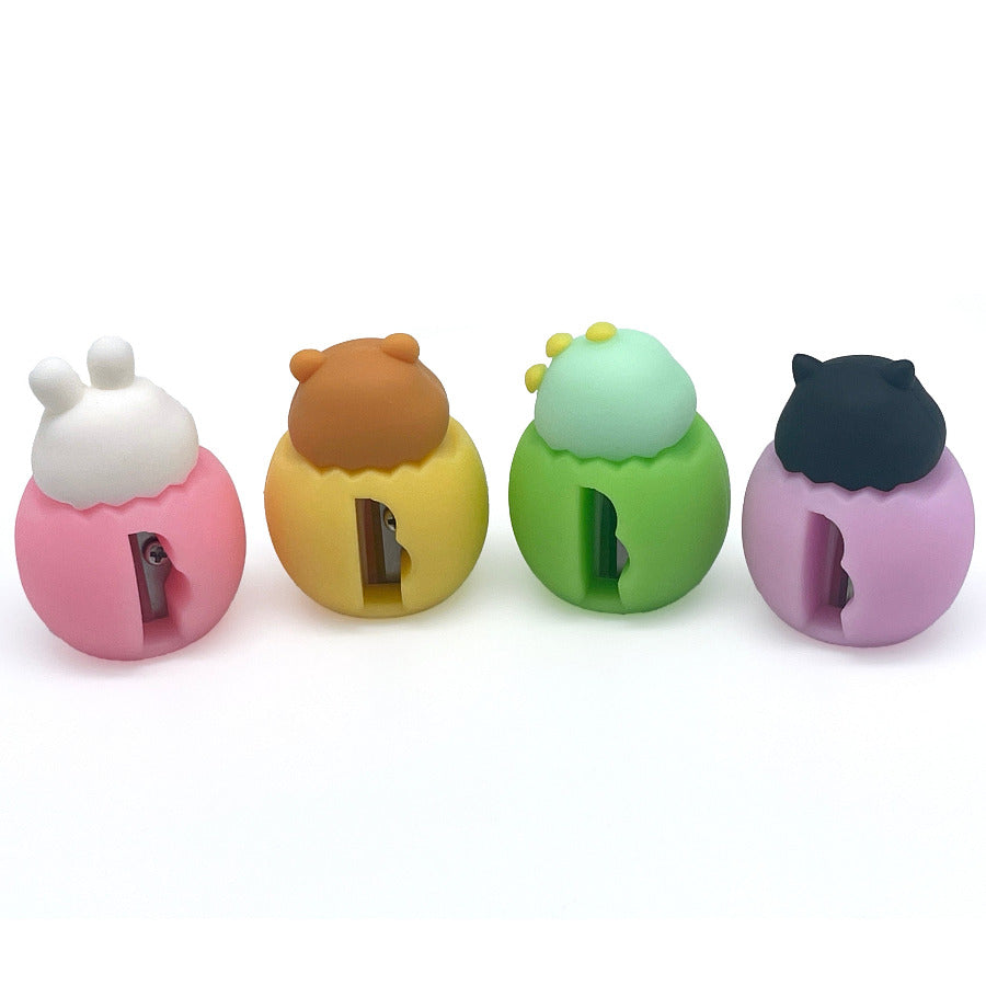 Four cute egg shaped pencil sharpeners with baby animal heads lined up in a row on a white background. From left to right, they have a pink rabbit head, a yellow bear head, a green dinosaur head, and a purple cat head.