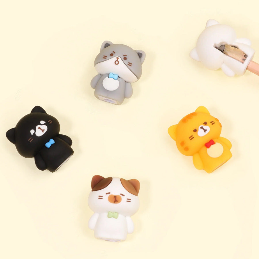 Five cute kawaii cat-shaped pencil sharpeners arranged on a pale cream background, including a black cat, gray cat, orange/yellow cat, white cat, and a white pencil sharpener partially visible at the top right.