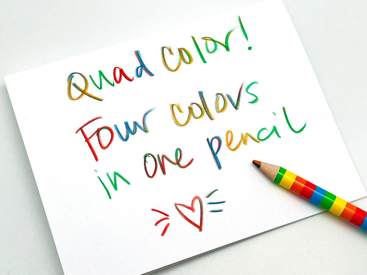 A white note card with colorful handwritten text reading 'Quad color! Four colors in one pencil' and a rainbow-striped colored pencil placed on the lower right, photographed on a light gray background.