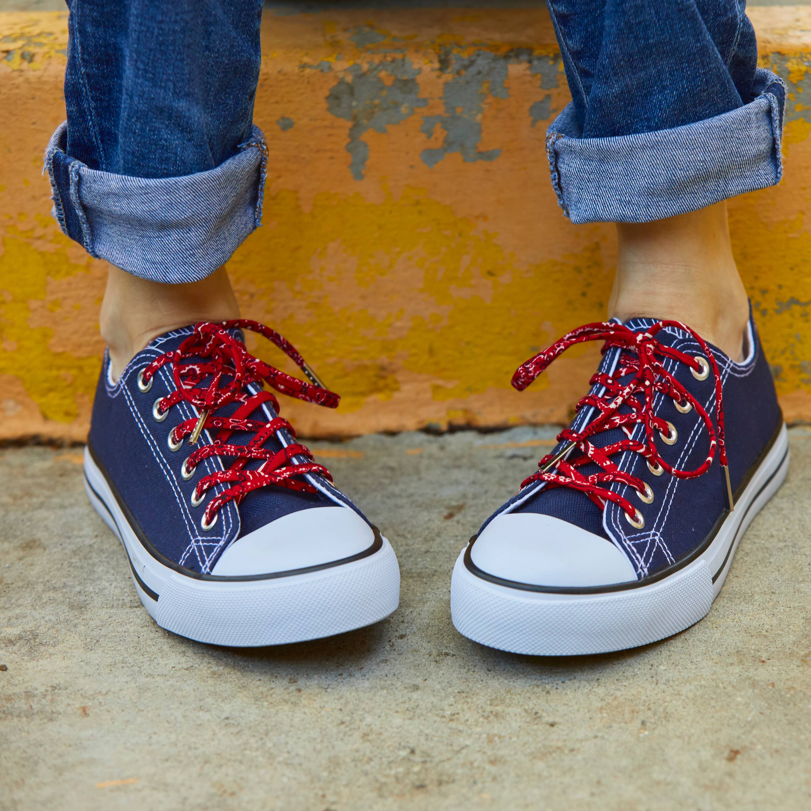 Navy sneakers with red laces worn with rolled-up jeans against a yellow wall.