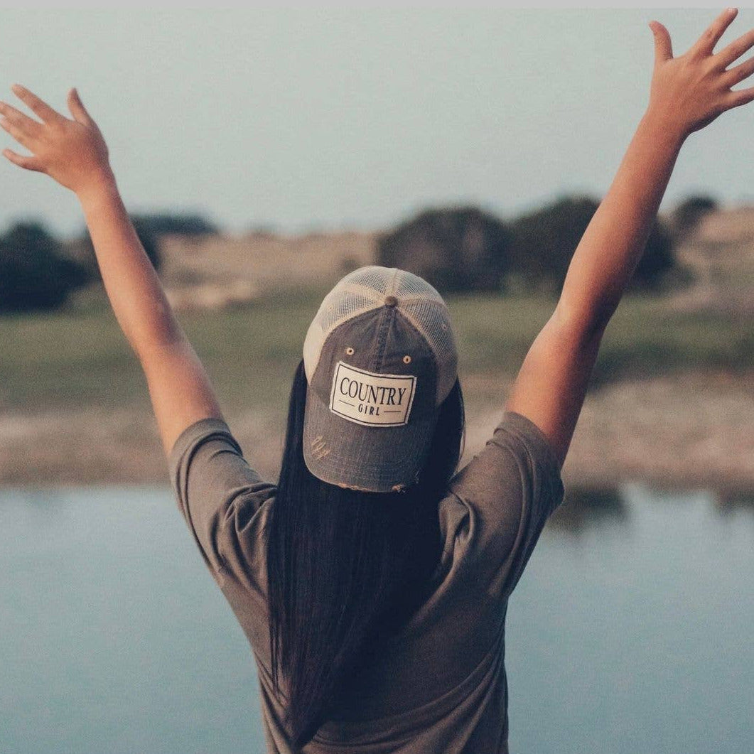 Person wearing a 'Country Girl' cap with arms raised by a body of water
