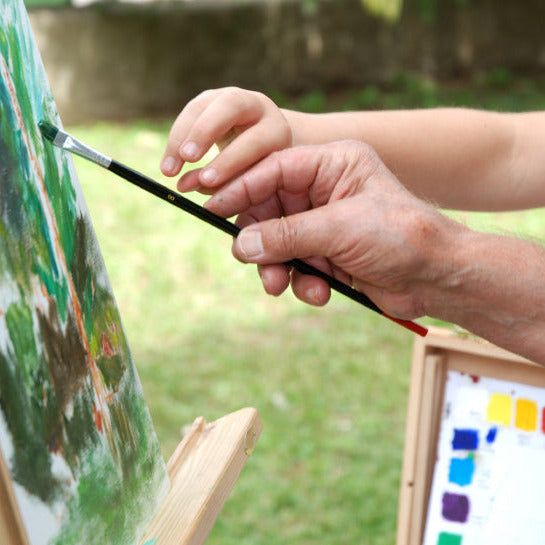 Adult guiding a child’s hand while painting outdoors on an easel promoting art supplies for kids and adults from Stubby Pencil Studio.