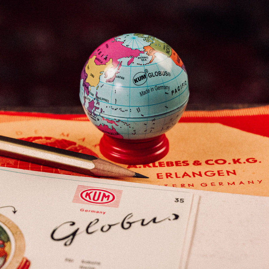 Small retro style world globe pencil sharpener on top of a KUM Globus box with a blurred background sitting next to a sharpened pencil.