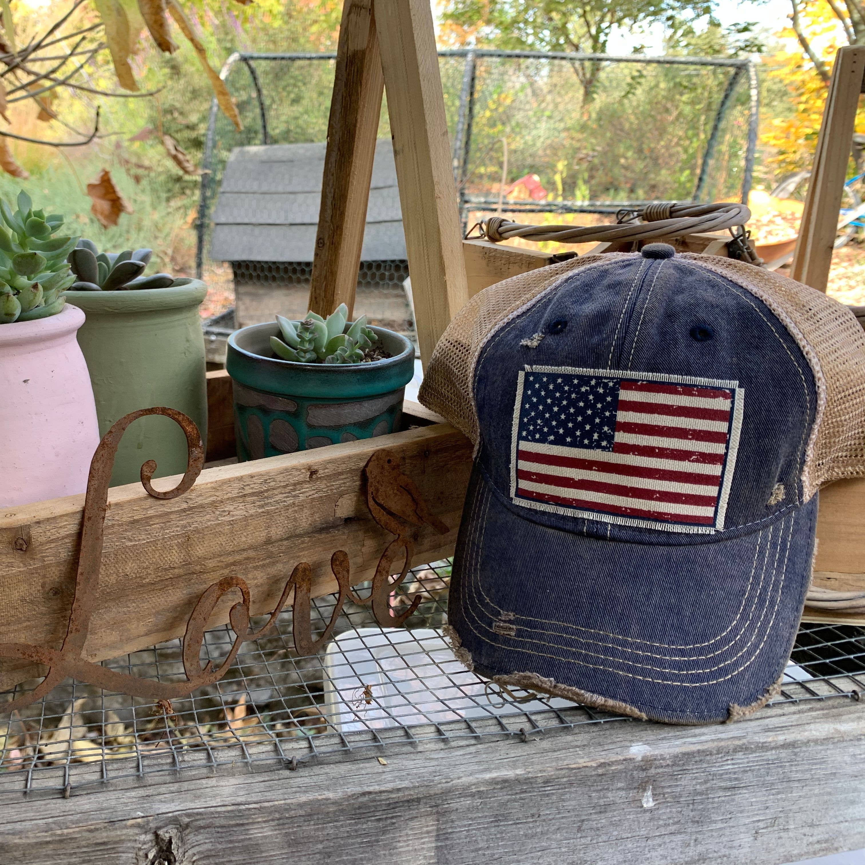 Blue cap with American flag patch on a wooden surface with potted plants in the background