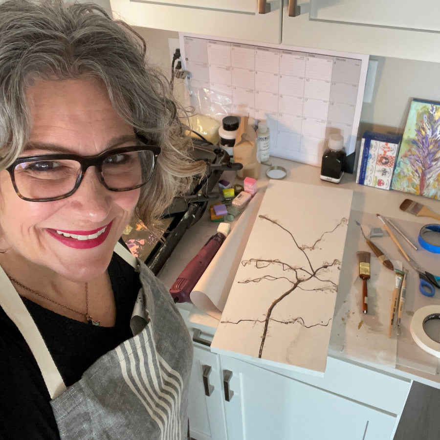 Female artist wearing an apron and standing next to an art panel with start of an encaustic painting with art supplies in the background