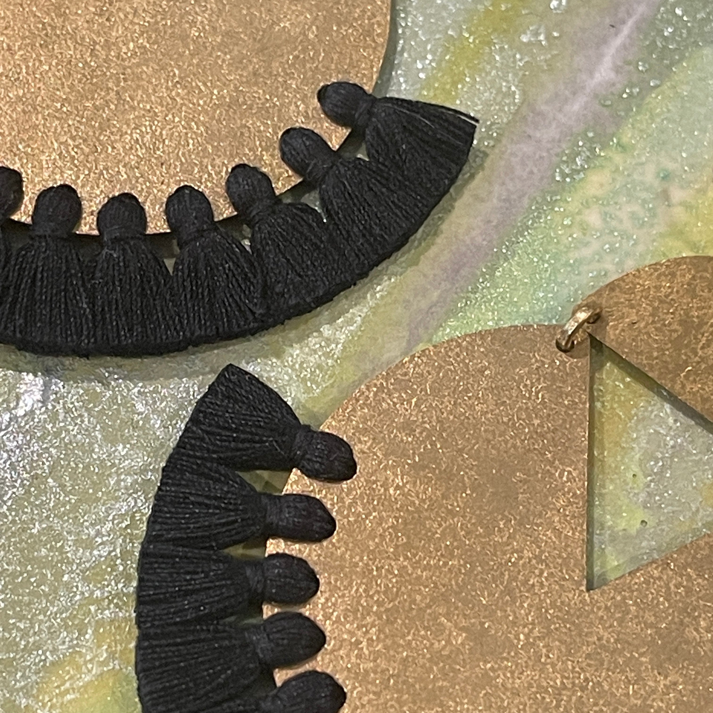 Close up detail of handmade brass dangle earrings with black color thread fringe detail on a metallic painting background