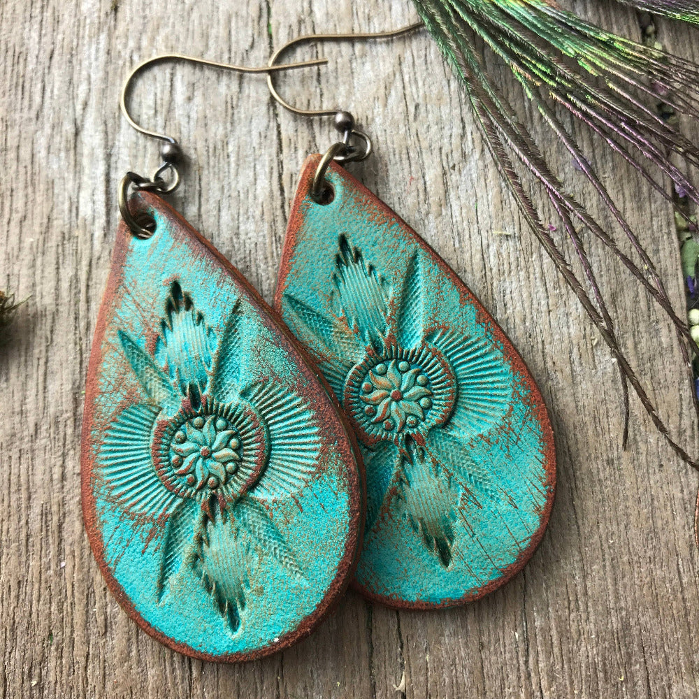 Handmade turquoise leather dangle earrings with embossed western boho hand tooled designs on a wooden surface with peacock feathers.