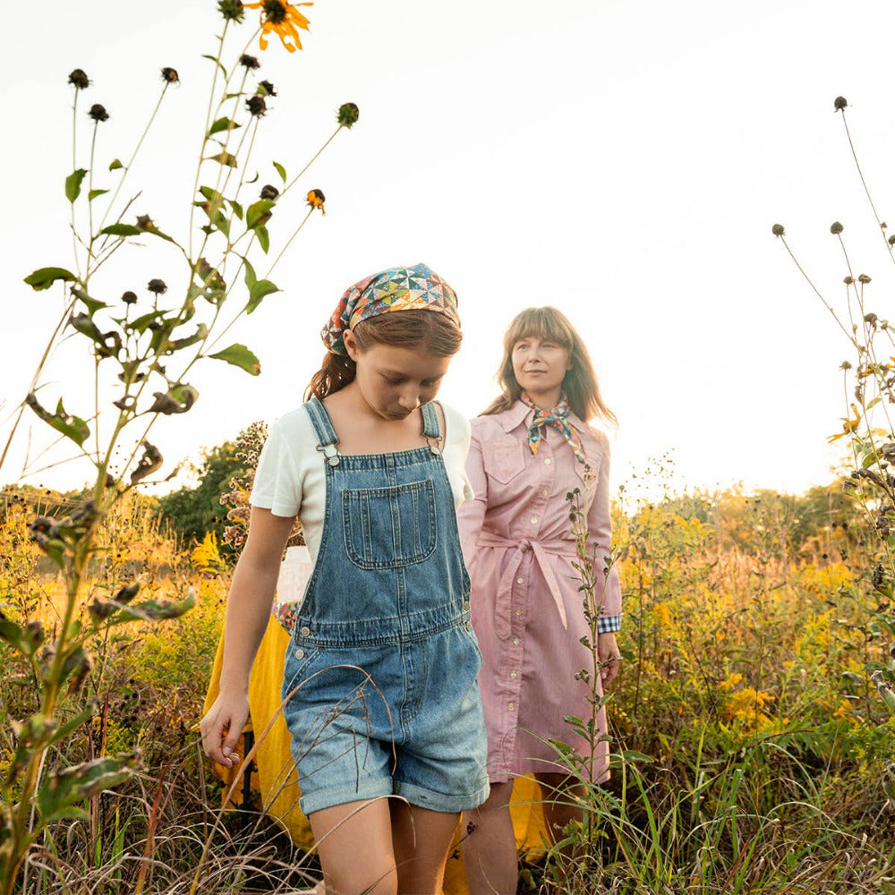 Two women walking outdoors in a sunlit field of wildflowers. One wears denim overall shorts over a white top with a colorful calico patterned cotton bandana as a head scarf, and the other wears a dusty pink dress with a matching neck scarf.