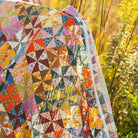 A colorful piece of fabric showing the Calico patchwork quilt pattern draped outdoors in a field of tall grass, featuring a geometric and floral pattern with many different colored triangles.