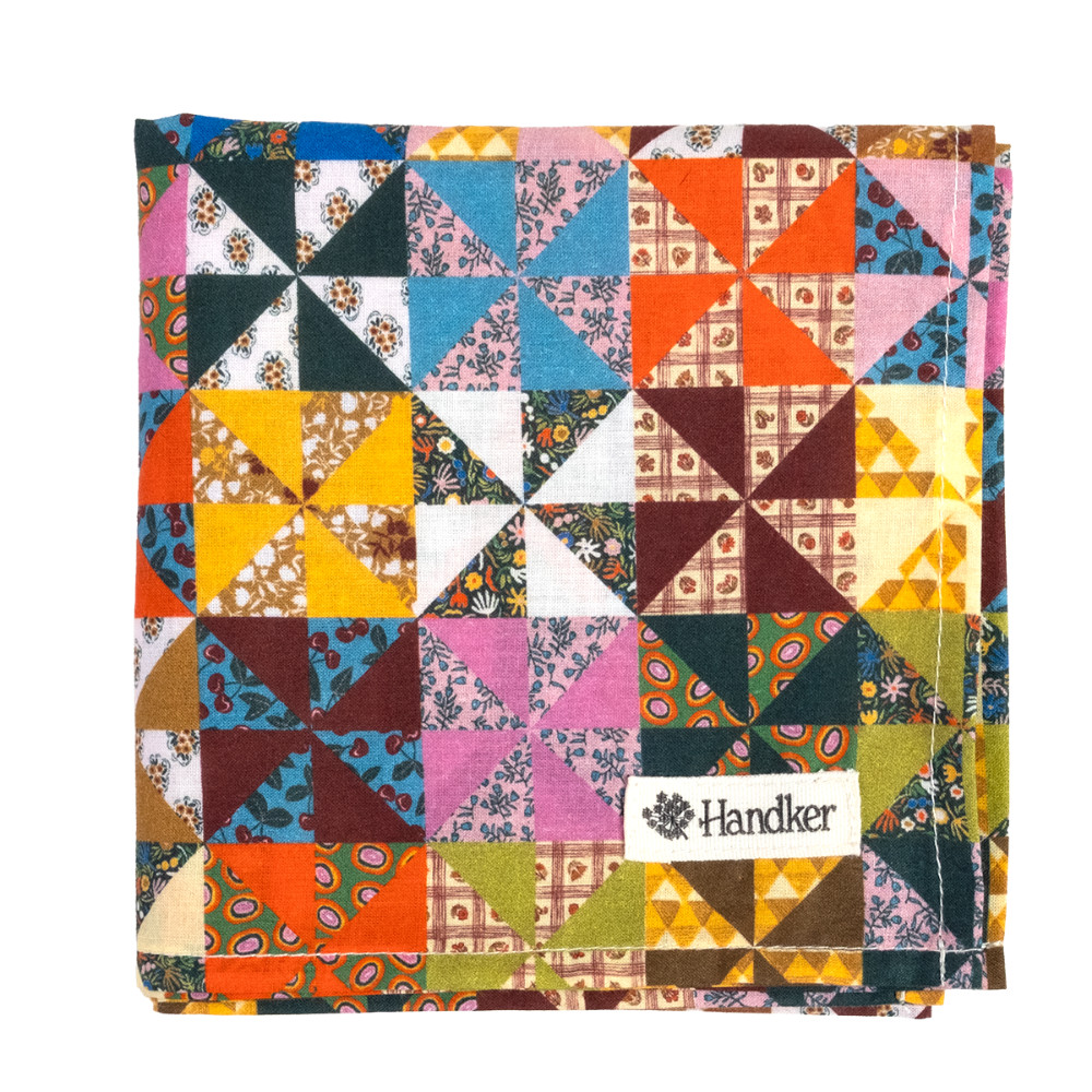 A folded square bandana featuring a colorful patchwork calico design with geometric and floral patterns, including orange, pink, blue, yellow, green, and brown. A white fabric label reading Handker is sewn onto the lower right area.
