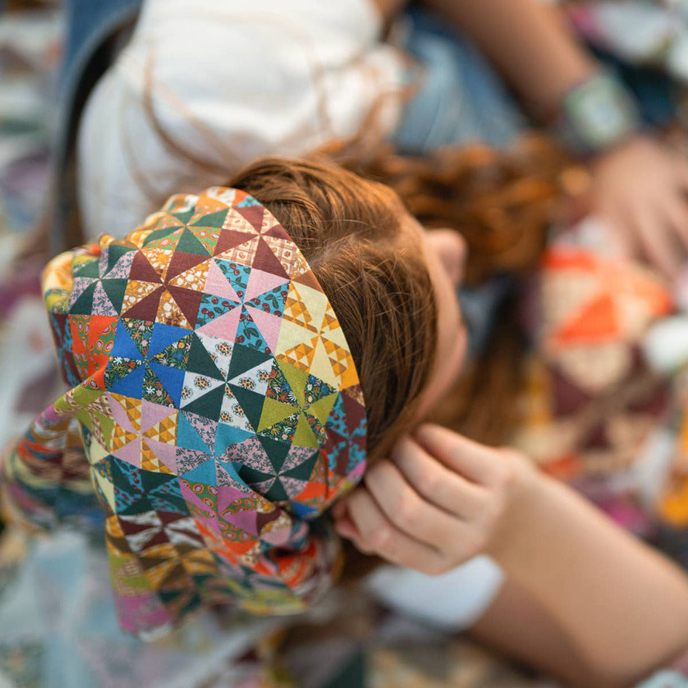 A close-up lifestyle shot of a person wearing a colorful Calico geometric patterned headscarf. The headscarf features a patchwork-style design with triangles in shades of blue, green, orange, pink, yellow, and brown.