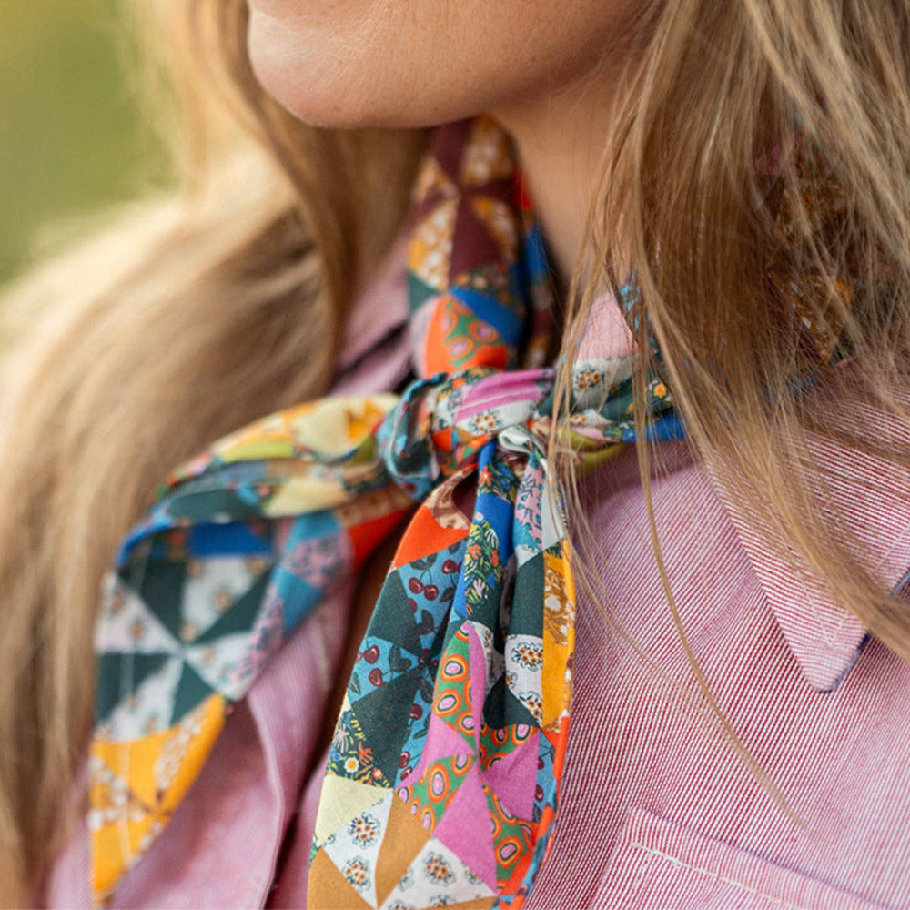 A close-up lifestyle shot of a woman wearing a vibrant, Calico multicolored patchwork-patterned cotton bandana scarf tied around her neck, paired with a pink striped button-down shirt.