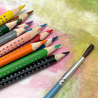 A set of grip watercolor pencils in various colors, each featuring a unique ergonomic triangular design with soft dots for grip and a paint brush on a colorful background.