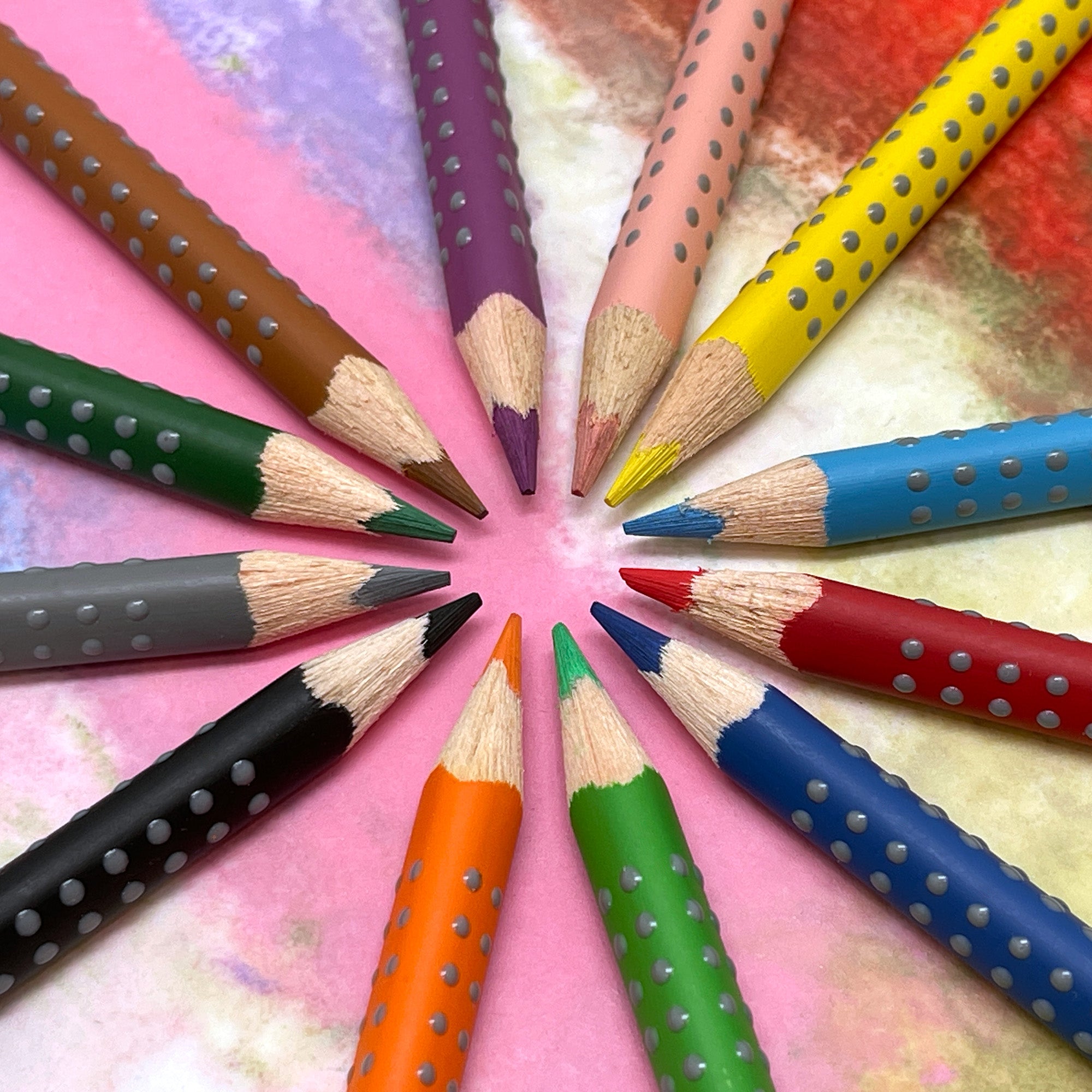 A set of grip watercolor pencils arranged in a circle in various colors, each featuring a unique ergonomic triangular design with soft dots for grip on a colorful background.