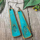 Turquoise leather earrings with brown accents on a wooden surface with green leaves.