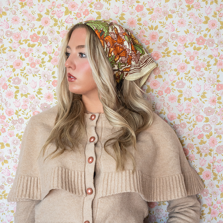 Woman wearing a beige cardigan and floral patterned headscarf against a floral wallpaper background