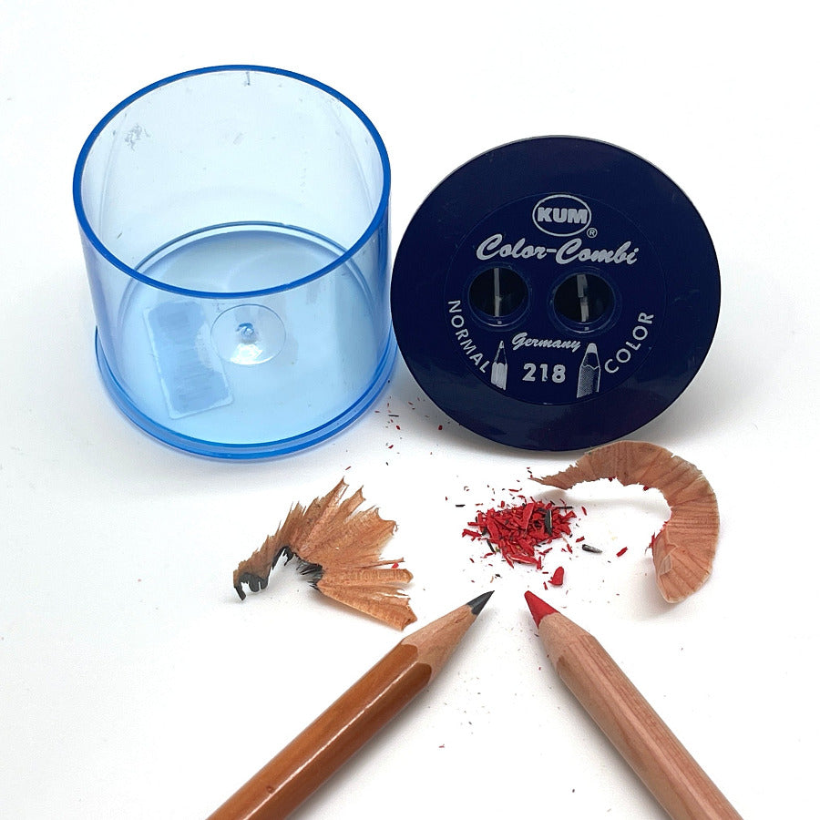 A KUM Color-Coni pencil sharpener set shown on a white background, including a transparent blue cylindrical container, a dark blue circular sharpening head with white printed branding, two wooden pencils, and shavings of wood and red pencil lead.