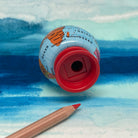 A mini globe-shaped pencil sharpener with a red circular base lies on its side on a blue watercolor background, with a wooden pencil placed diagonally in the lower left corner.