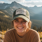 Person wearing a cap with 'Mountain Girl' text against a mountainous landscape