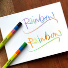 Jolly Rainbow pencils set on a white note card with the words "Rainbow" written twice showing the rainbow swirl writing and a drawn line below set on a wooden table